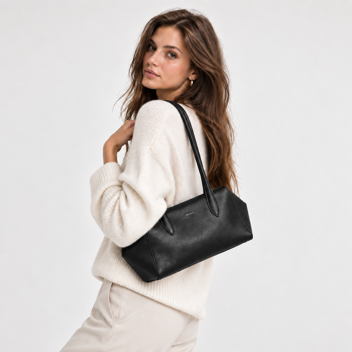 A woman wearing a cream knit sweater and light beige pants carries a black leather shoulder bag against a plain light gray studio background.