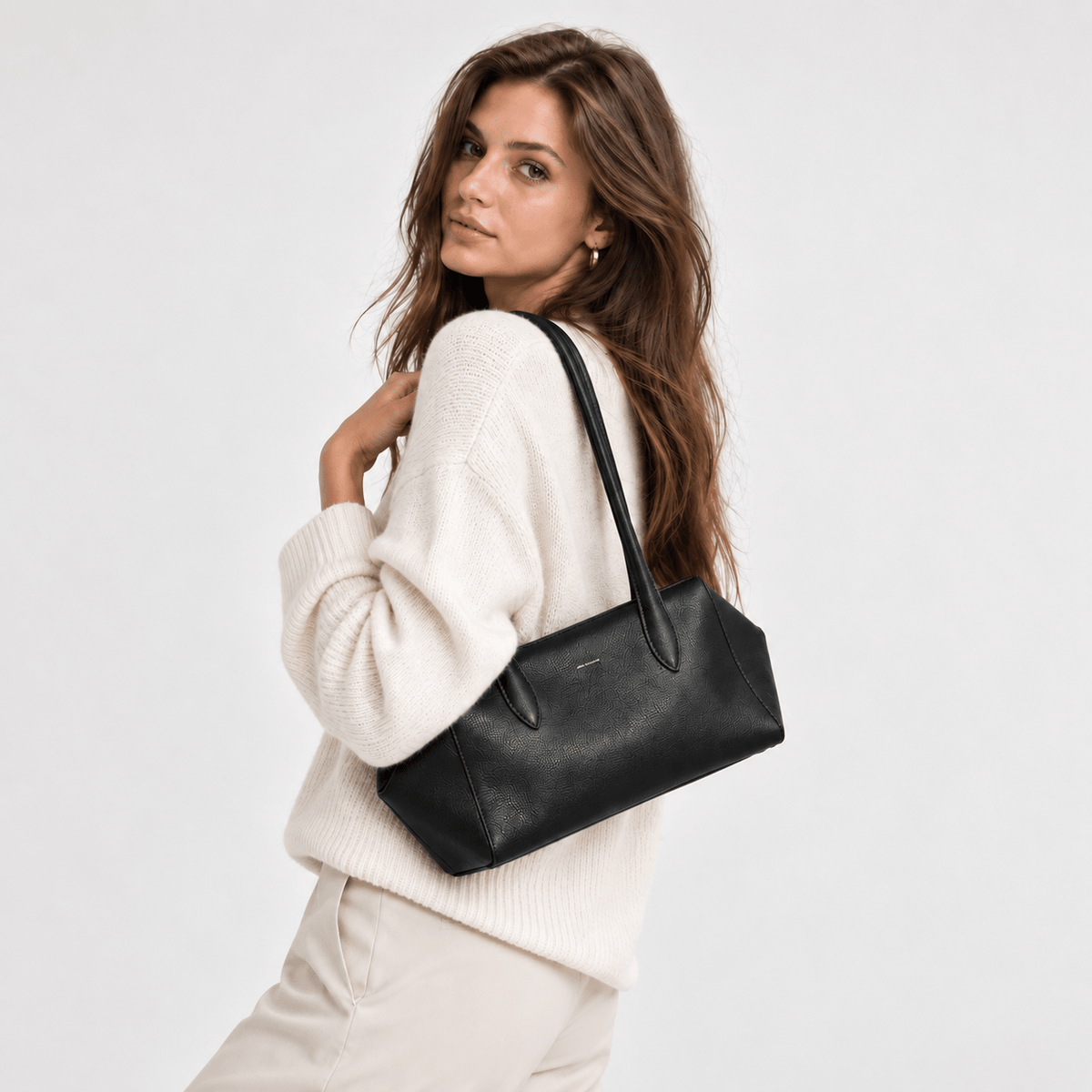 A woman wearing a cream knit sweater and light beige pants carries a black leather shoulder bag against a plain light gray studio background.