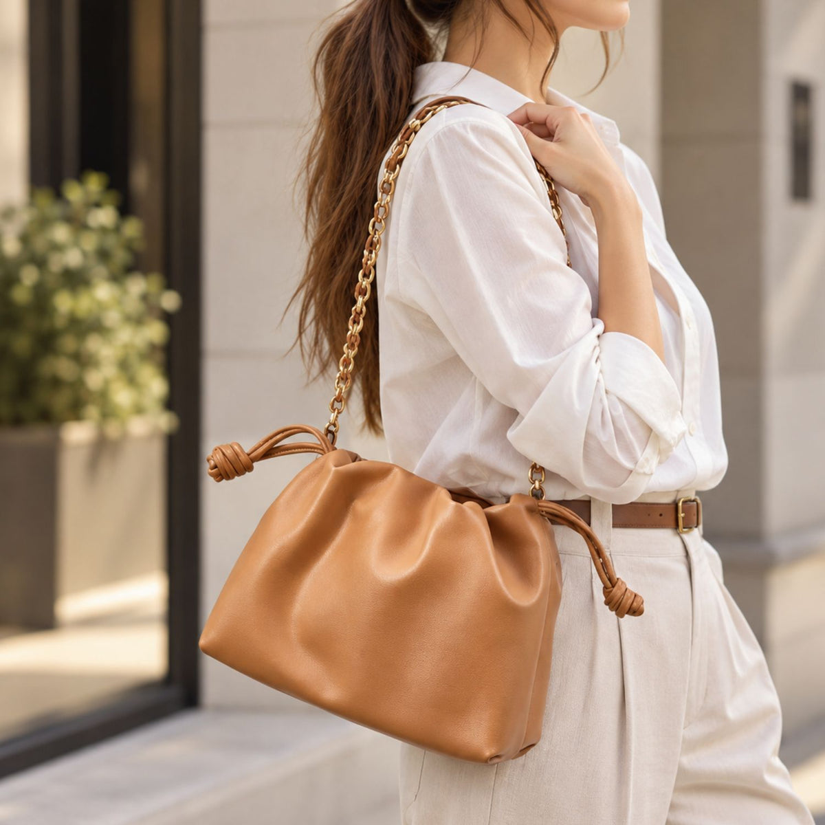 A woman in a white shirt and light pants carries a tan leather slouchy shoulder bag with a gold chain strap and knotted leather handles, photographed in an outdoor urban setting.