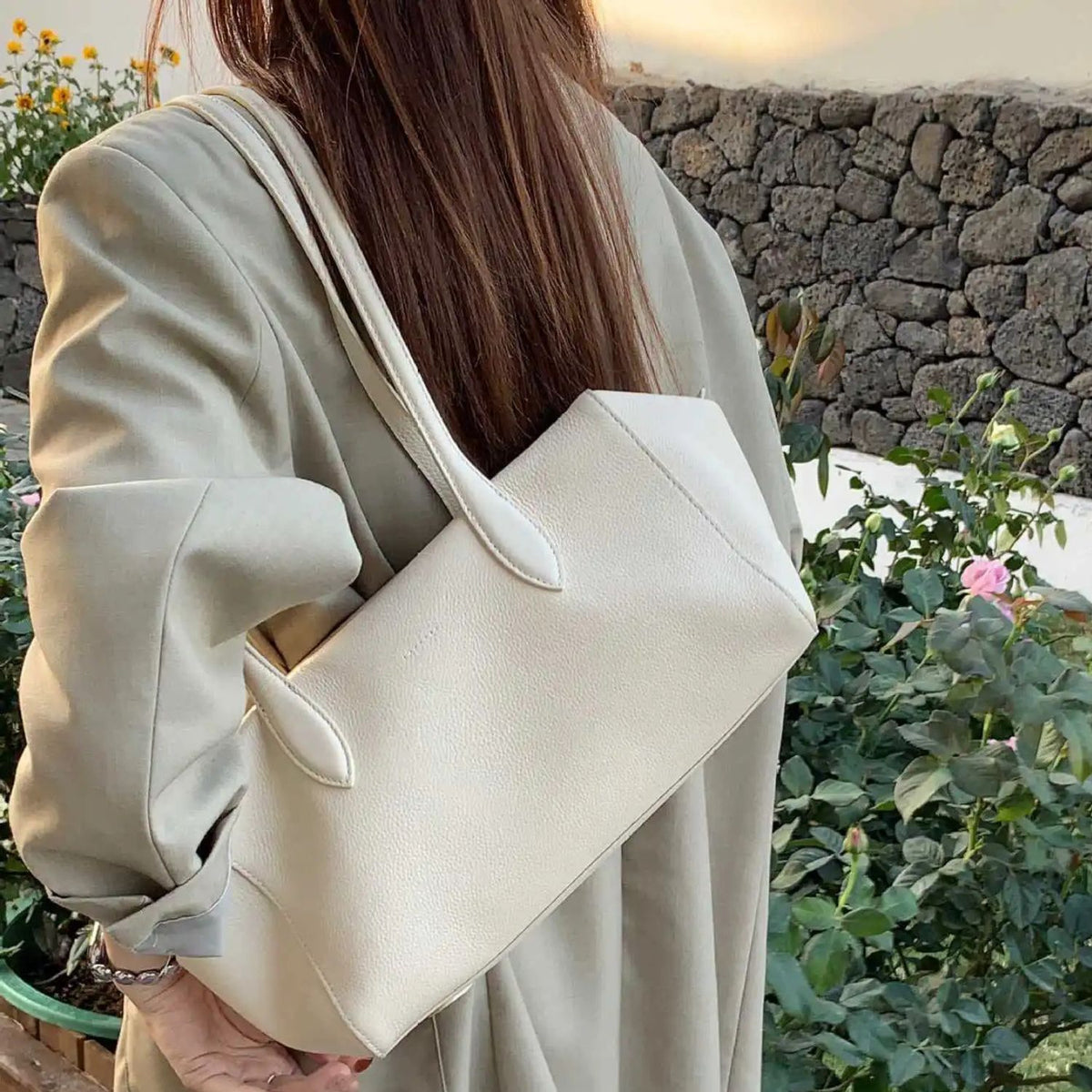 A woman wearing a light beige blazer carries a large, structured off-white tote bag over her shoulder in an outdoor garden setting with a stone wall background.