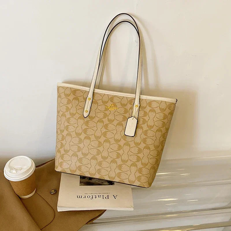 Beige handbag with brand logo on a white surface with a coffee cup and magazine.