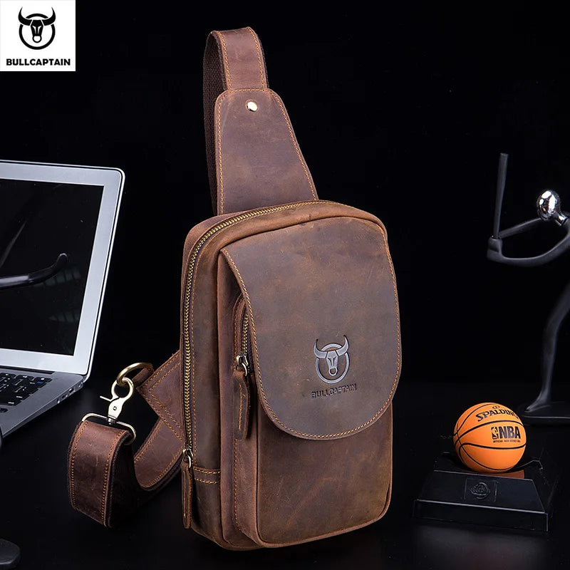 Brown leather sling bag with Bullcaptain logo on a dark surface with laptop and basketball.