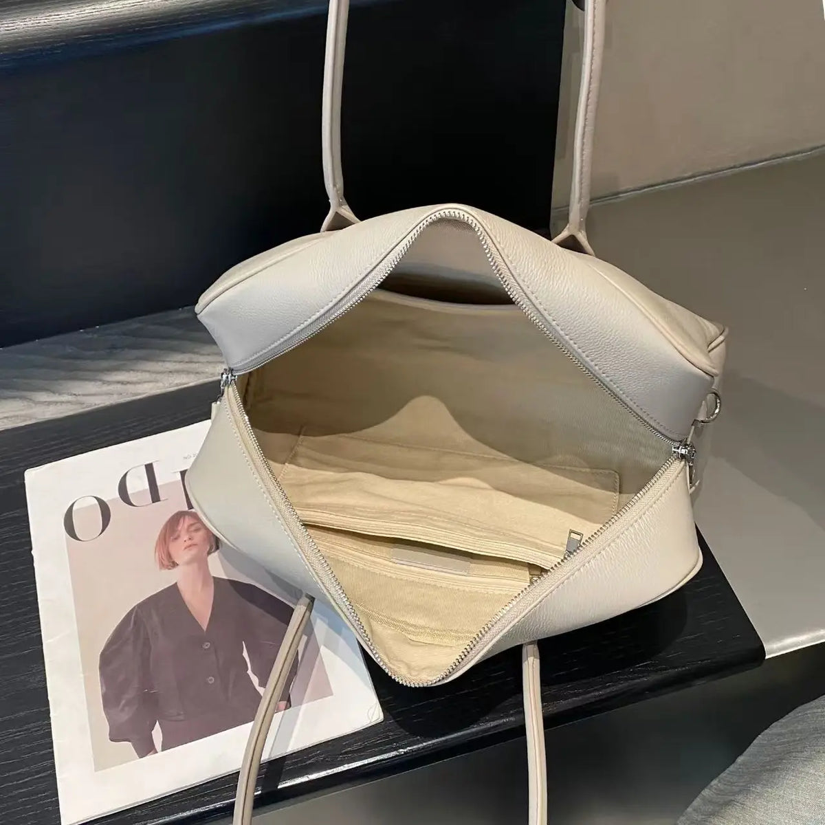 A light beige faux-leather shoulder bag with a zippered main compartment and interior organizer pockets, shown open on a dark table next to a magazine.