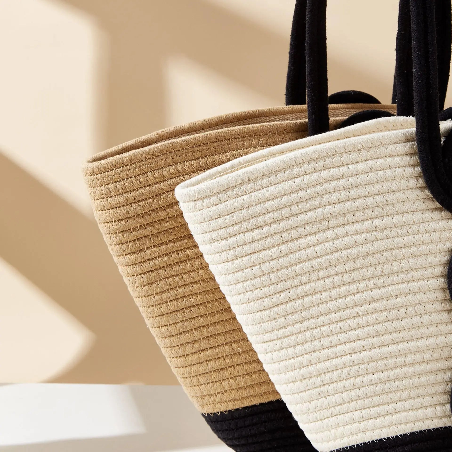 Two woven bags, one beige and one white, with black handles on a neutral background.
