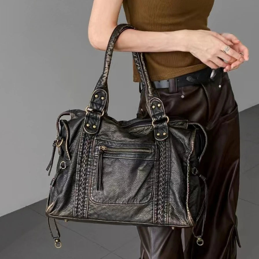 Woman wearing brown leather pants and a sleeveless top, holding a black Y2K leather shoulder bag with braided details and metal hardware.