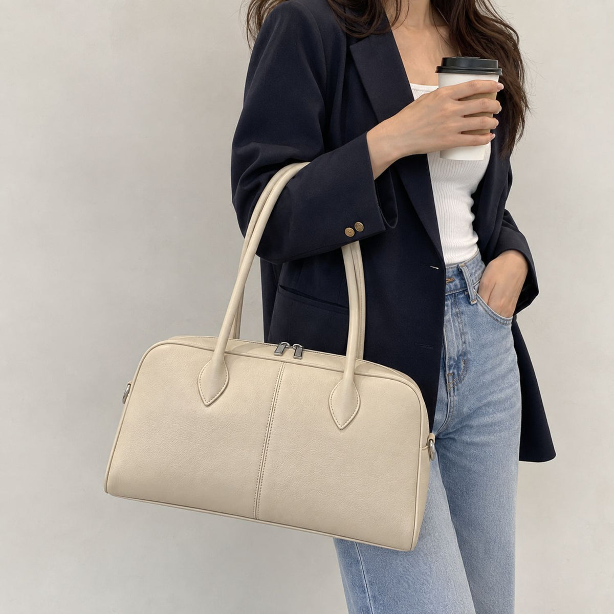 A woman wearing a navy blazer, white top, and light blue jeans carries a large cream-colored leather duffel bag with double top handles while holding a black coffee cup against a plain light gray wall.