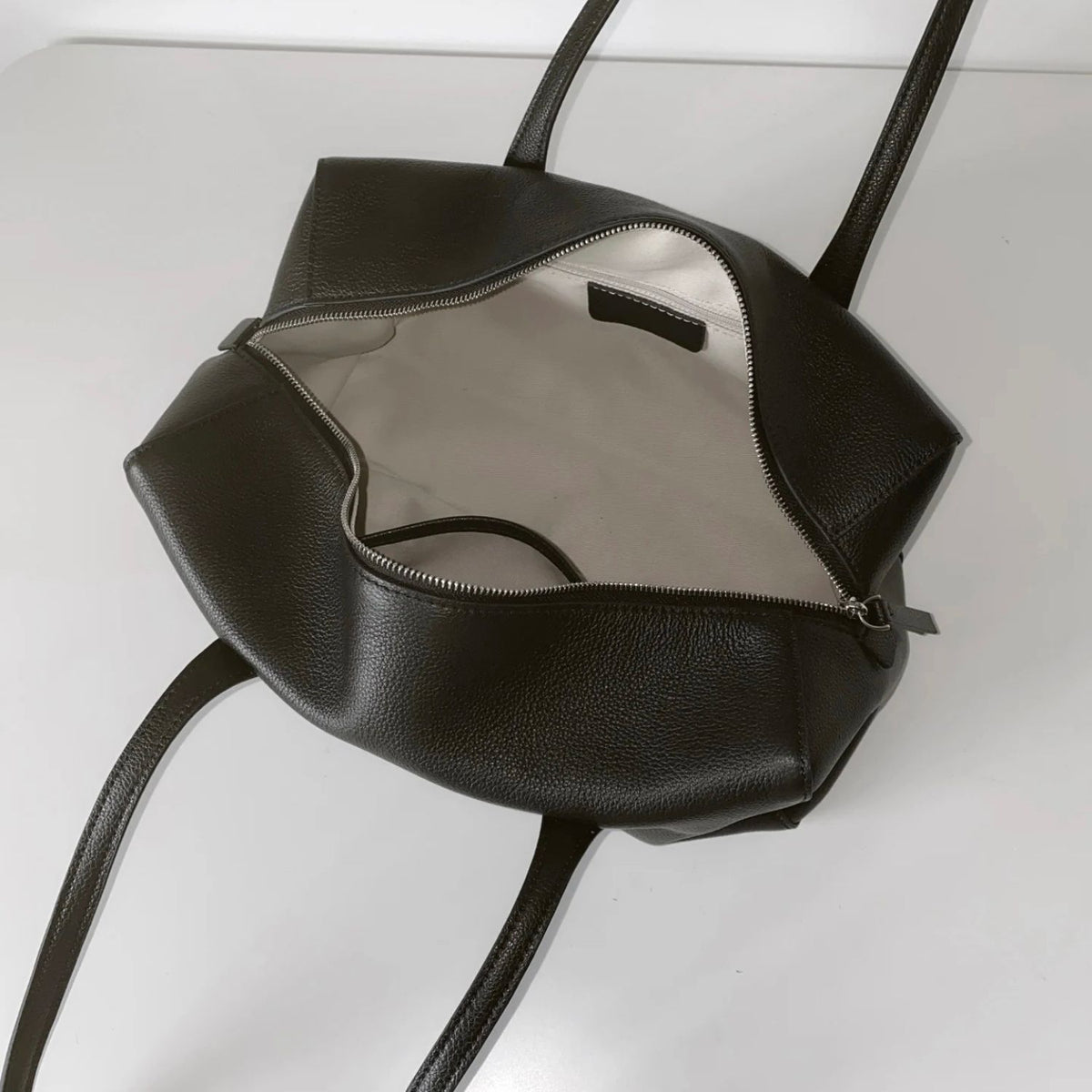 A black pebbled leather handbag shown open from a top-down angle, revealing a light beige interior lining and a silver zipper. The bag has a rounded, slouchy shape with long shoulder straps and is photographed on a clean white surface.