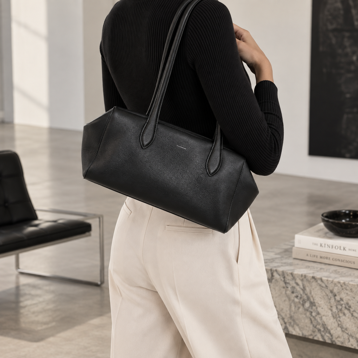 A person wearing a black ribbed long-sleeve top and cream wide-leg trousers carries a large black leather shoulder bag with a structured rectangular shape and long handles in a modern indoor setting.