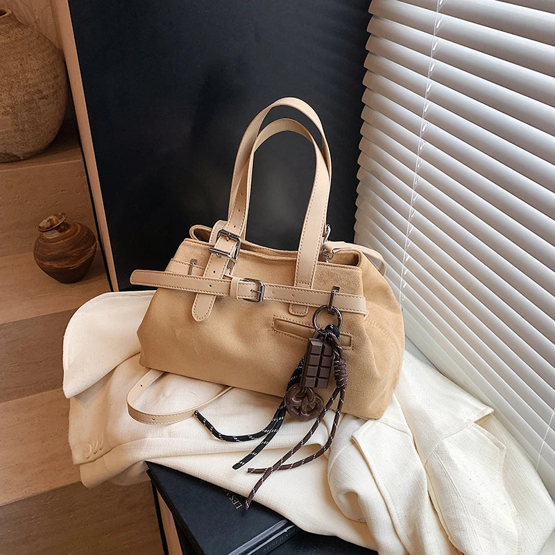Beige handbag with decorative keychain on a white surface near a window with blinds.