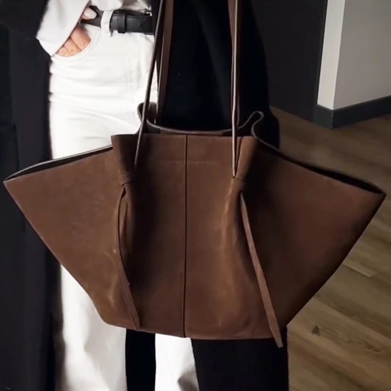 Brown leather tote bag held by a person wearing a black coat and white pants.