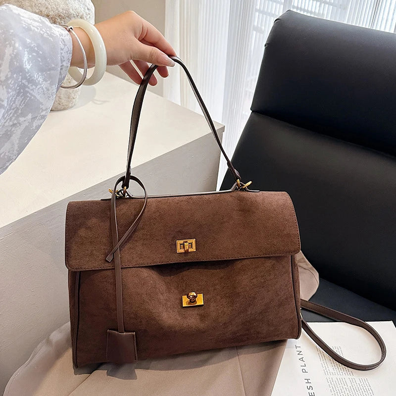Brown suede handbag with gold clasp held by a person indoors.