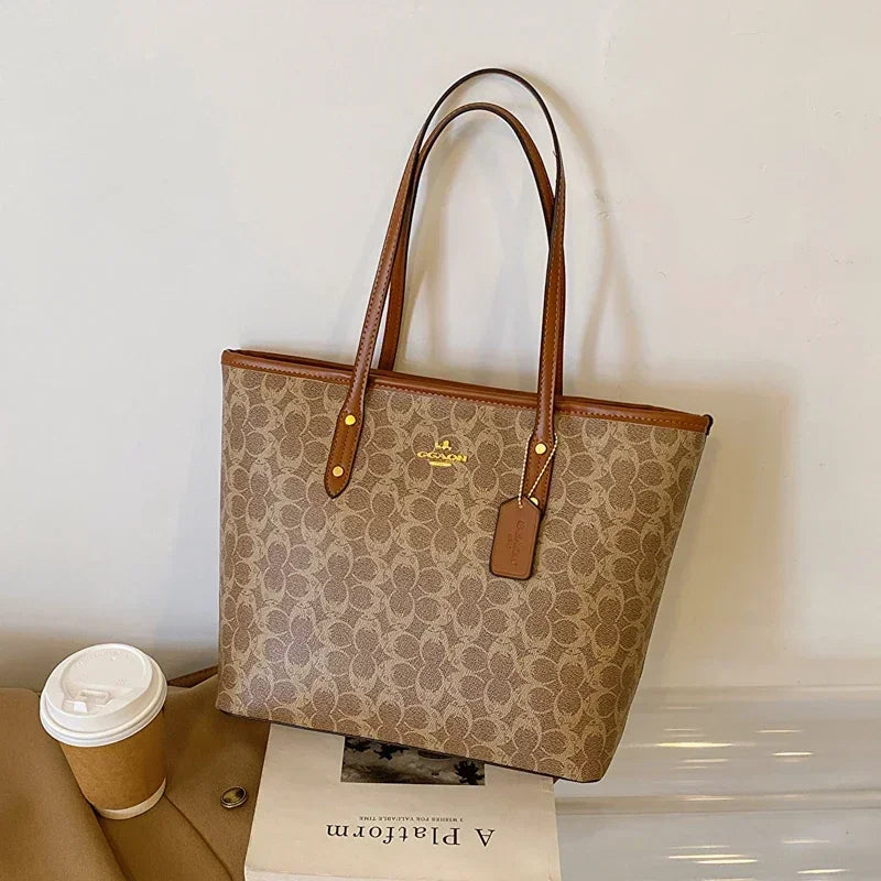 Coach handbag with brown handles on a white surface next to a coffee cup and book.