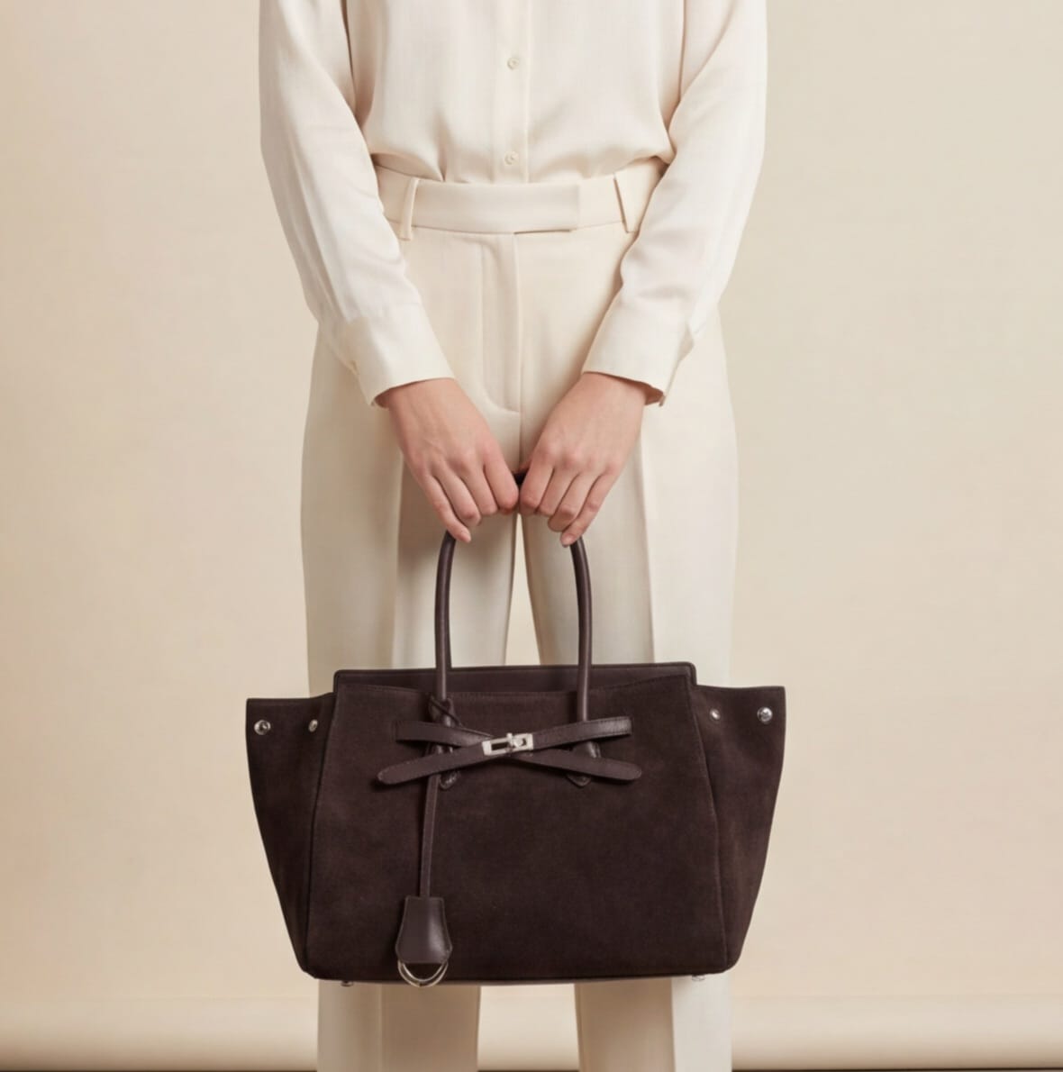 woman in cream outfit holding dark brown suede tote bag with lock detail by the handles