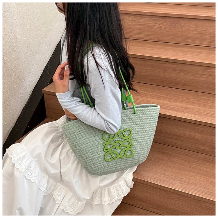 Person holding a green textured handbag with a logo, wearing a white outfit, against a wooden background.