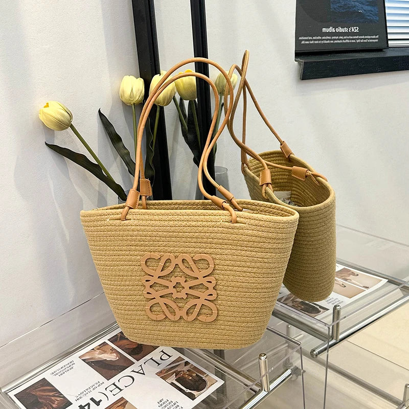 Two woven handbags with a visible brand logo on a glass surface with flowers and magazines in the background.