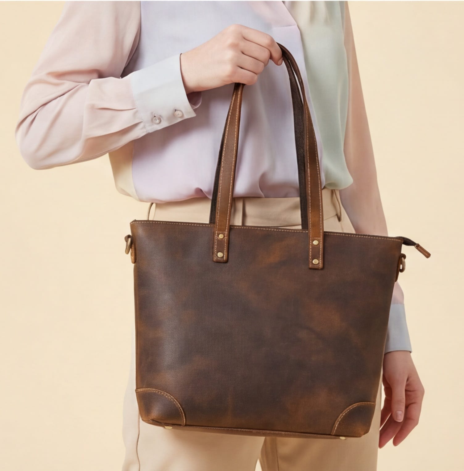 Brown leather tote bag held by a person against a beige background