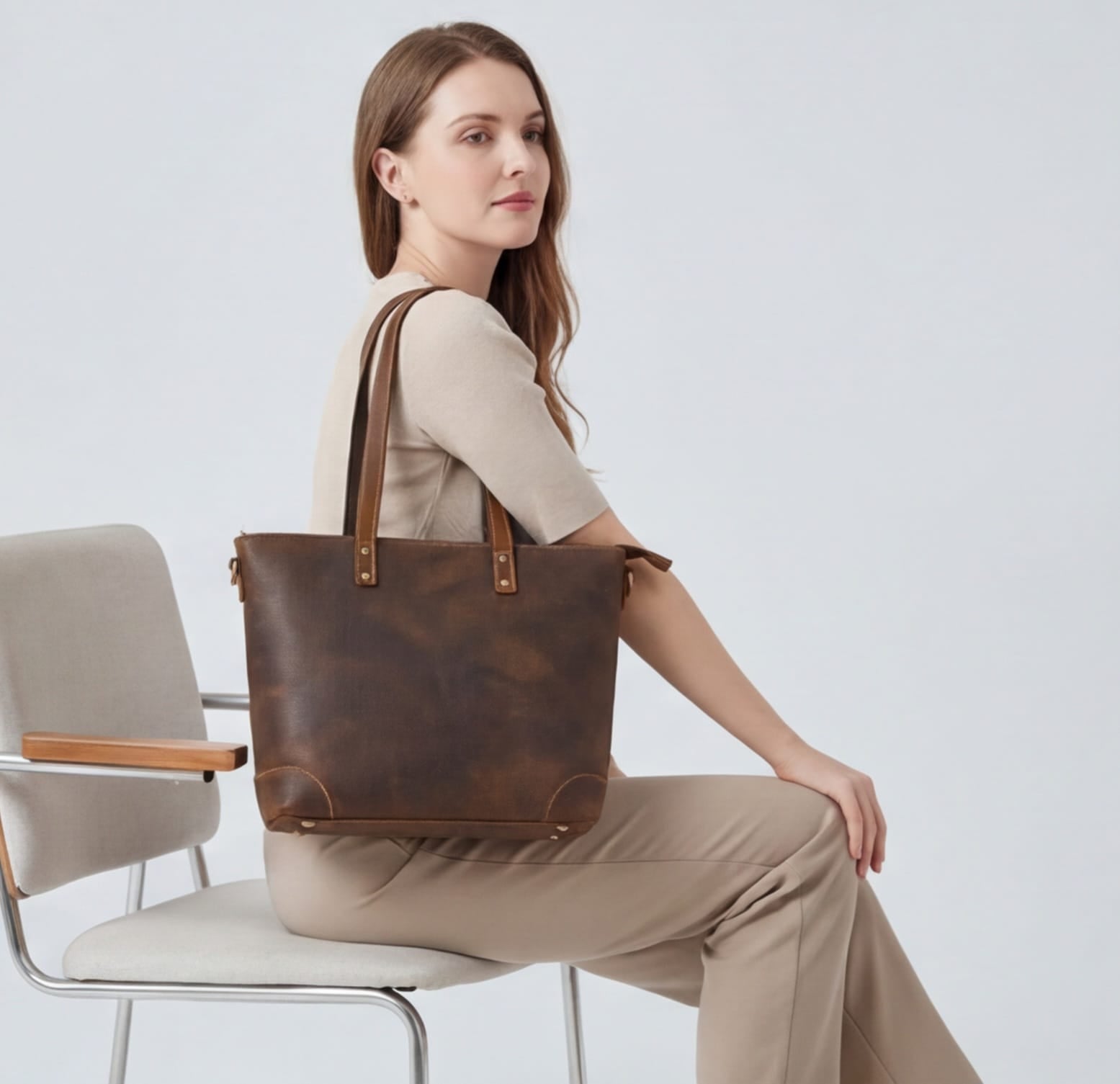 Woman sitting on a chair holding a brown leather tote bag against a plain background