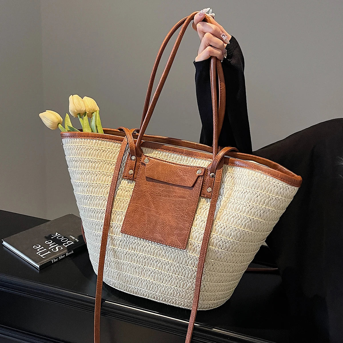 Woven handbag with brown leather accents held by a person, with tulips inside on a dark surface.
