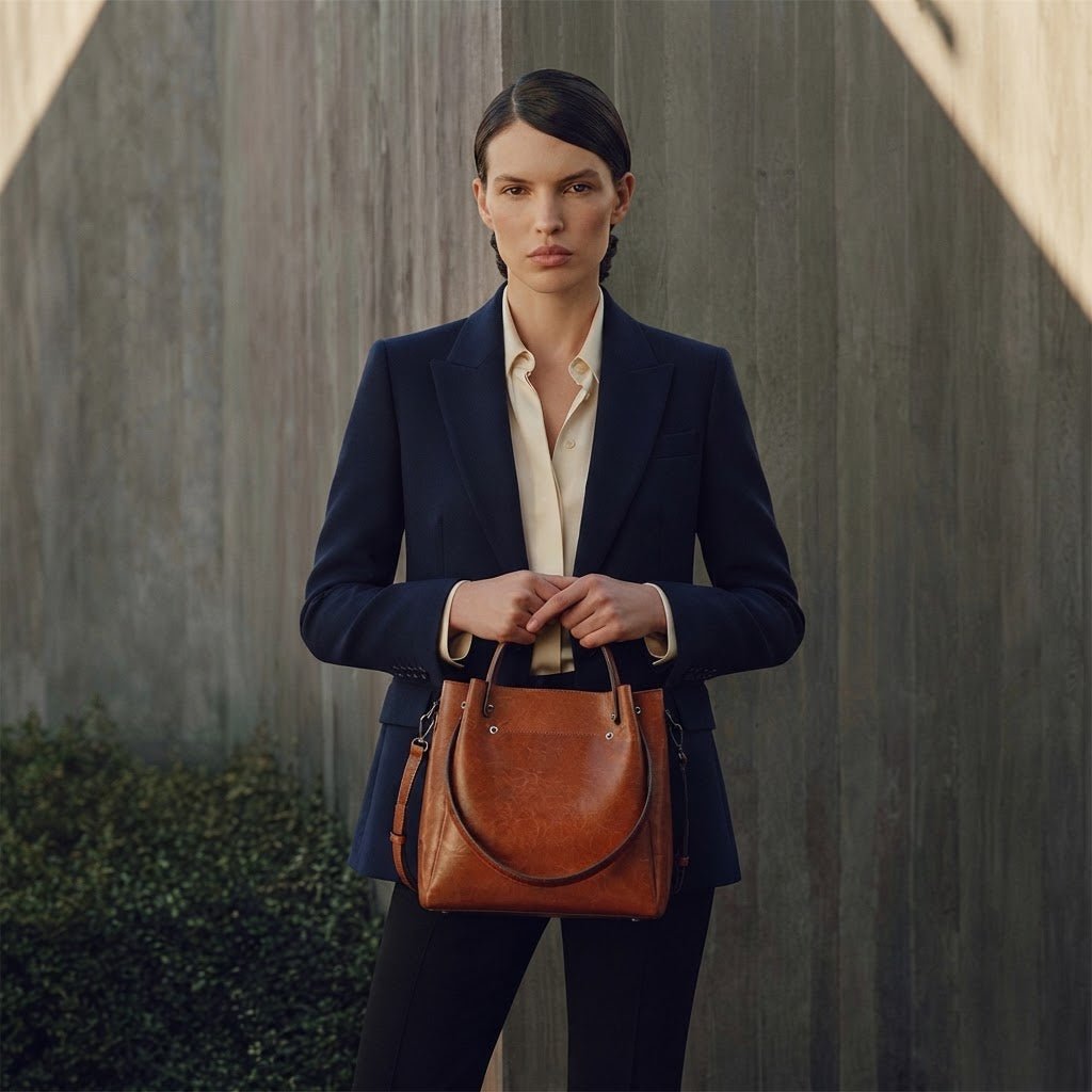 Woman wearing a navy blue tailored suit and cream blouse, holding the Amelia brown leather handbag with top handles and a shoulder strap against a modern concrete wall.