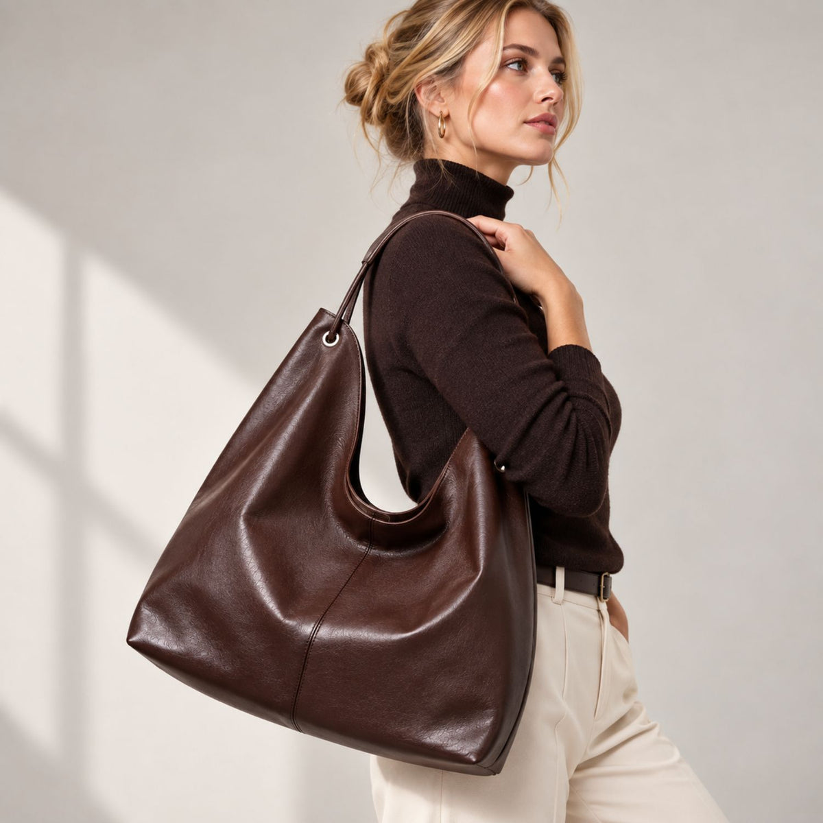 A woman wearing a dark brown turtleneck sweater and cream trousers carries a large, slouchy dark brown leather hobo shoulder bag against a light gray background with soft window shadows.