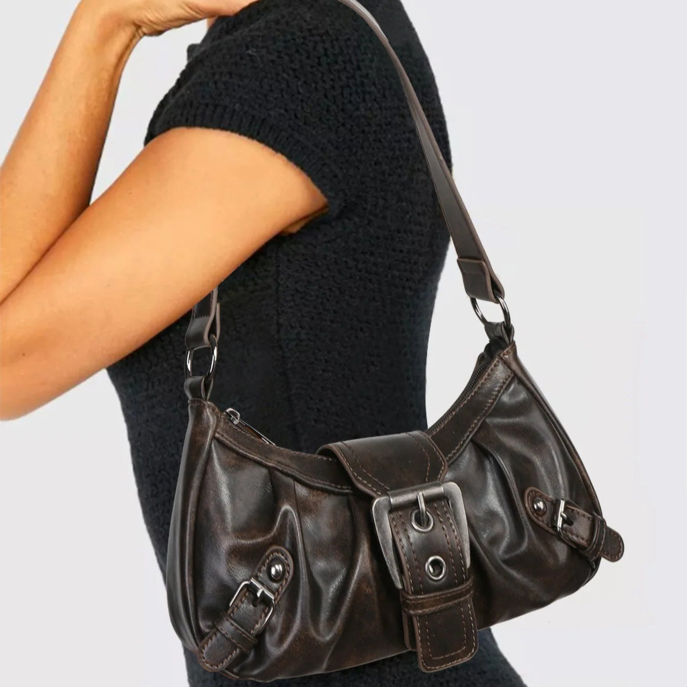 Woman wearing a black short-sleeve top holding a dark brown leather shoulder bag with buckle details.